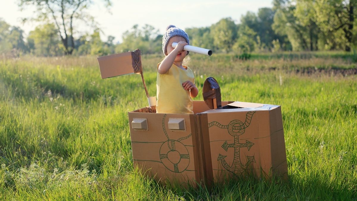 young child in cardboard boat outside looking through cardboard telescope