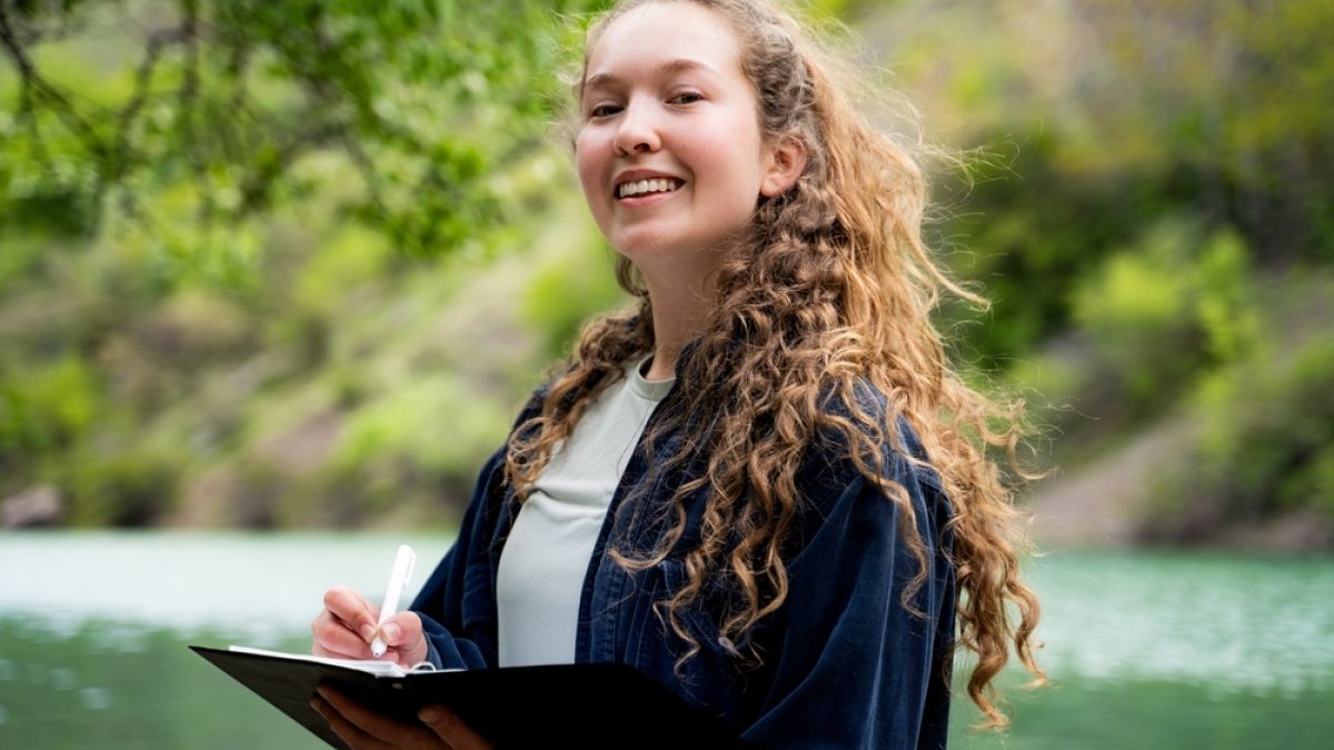 girl with long curly hair outside in nature with a pen and notebook