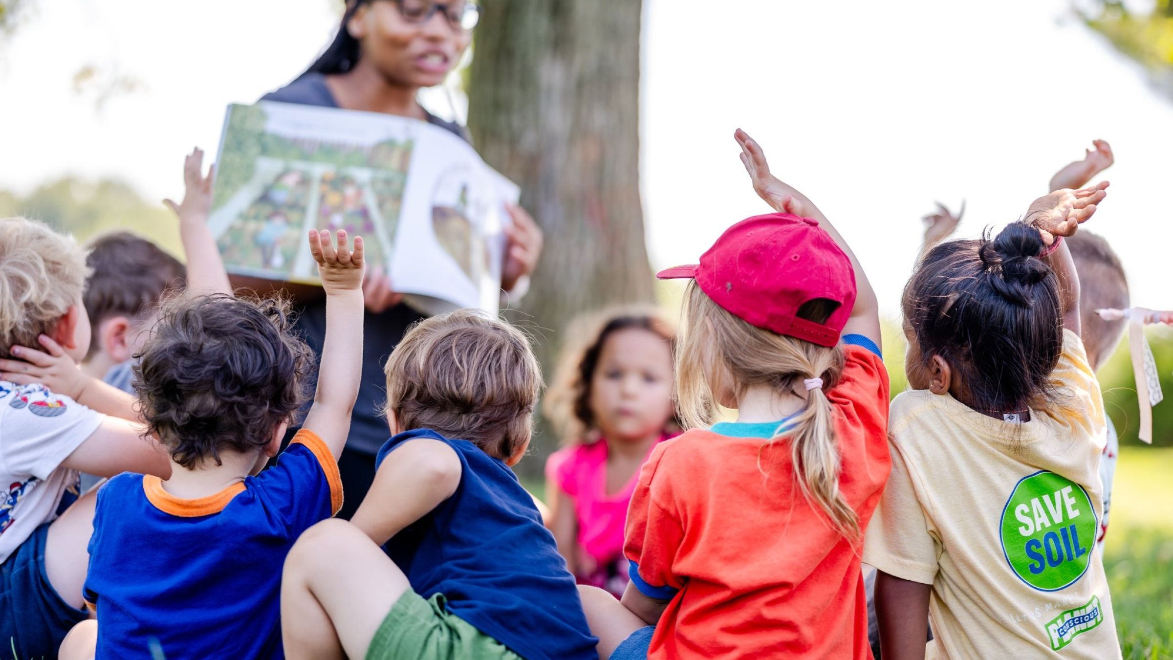 children sitting outside with a teacher raising their hands to be called on