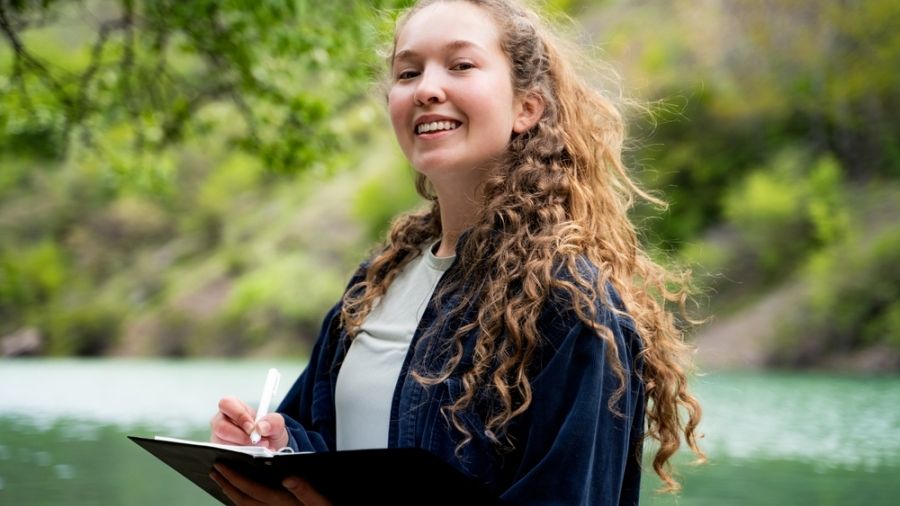 girl with long curly hair outside in nature with a pen and notebook