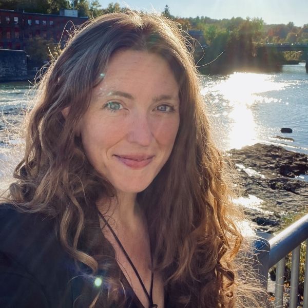 Image of a woman with curly red hair smiling against a backdrop of a river with the sun shining