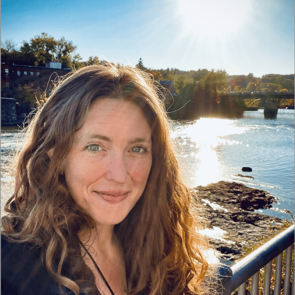 Image of a person with long reddish hair standing by a river and smiling at the camera