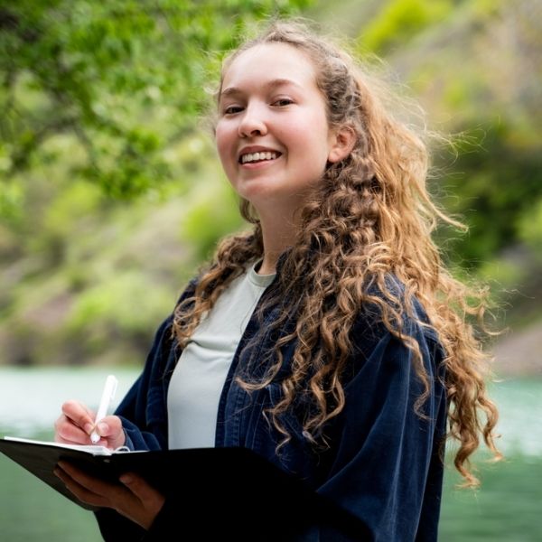 girl with long curly hair outside in nature with a pen and notebook