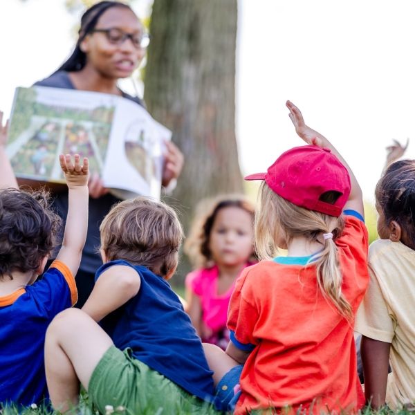 children sitting outside with a teacher raising their hands to be called on